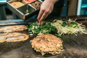 man chef hands adding green onion vegetable in cooking okonomiyaki in local restaurant on teppanyaki pan. delicious tasty japanese style pancake street food on iron plate hot. noodles under eggs